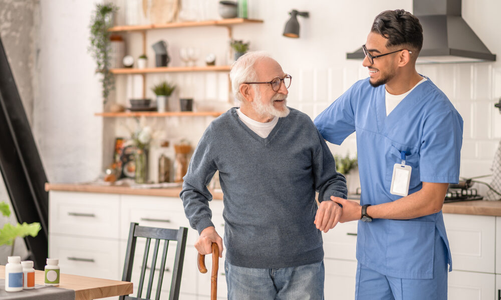 Smiling friendly in-home male nurse in uniform supporting an old man with a walking stick